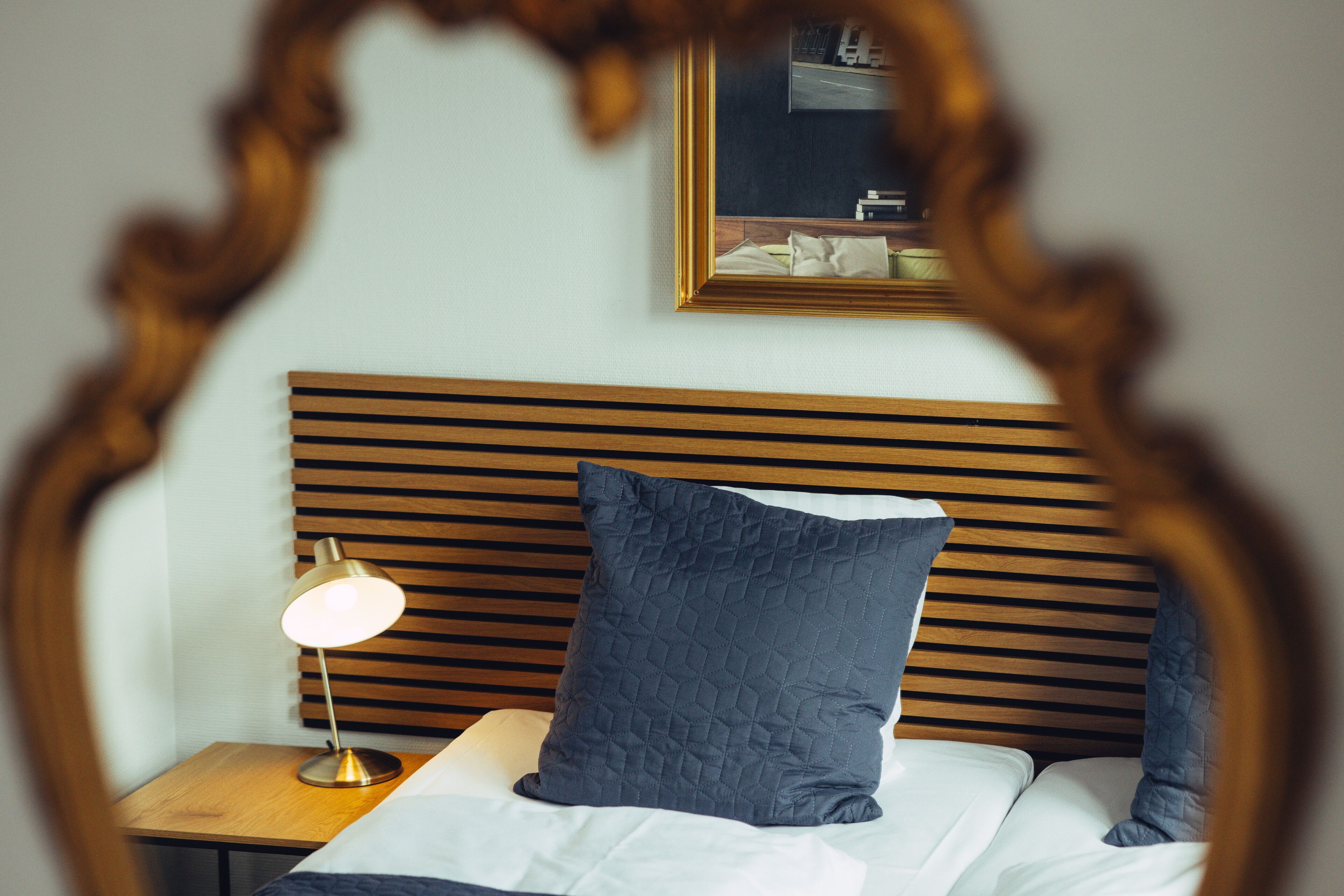 A neatly made bed with a wooden headboard, a bedside lamp, and a framed mirror on the wall.