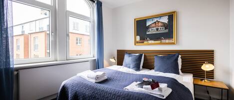Una habitación de hotel con una cama grande, un cabecero de listones horizontales, una ventana con cortinas, un cuadro colgado en la pared y una mesita de noche con lámpara.