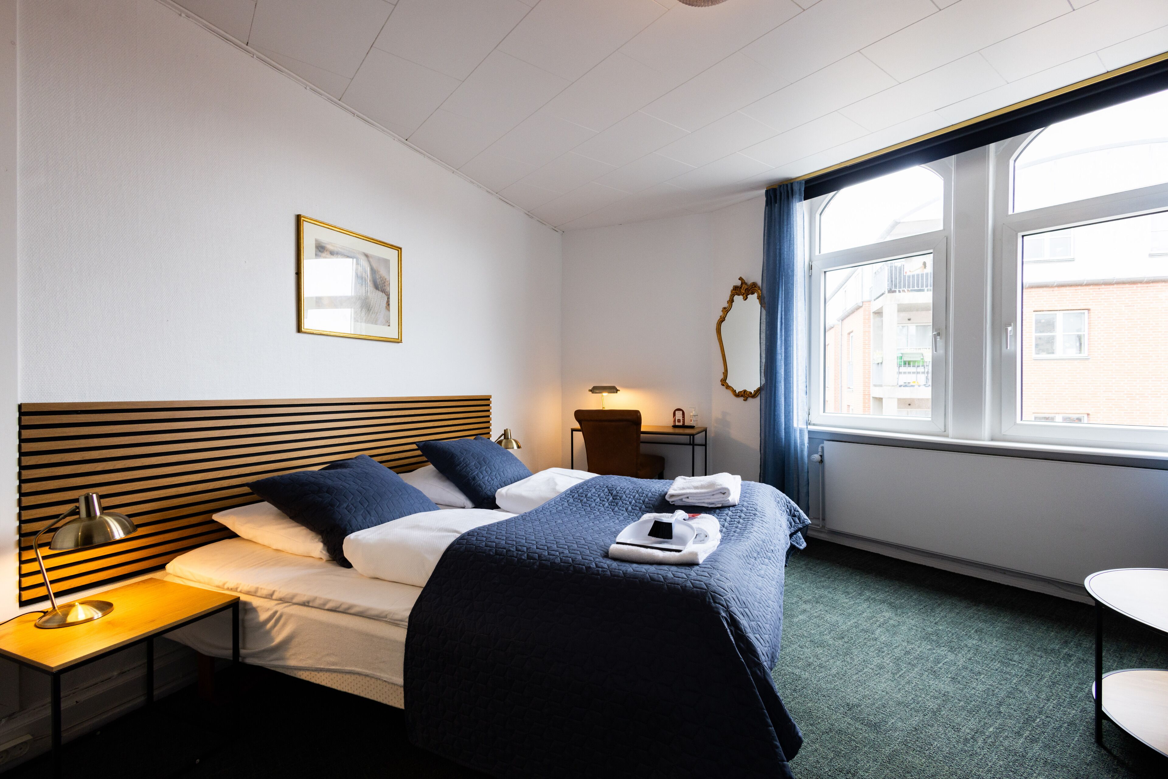 Habitación de hotel con cama, mesita de noche, lámpara y ventana con vista a edificios.