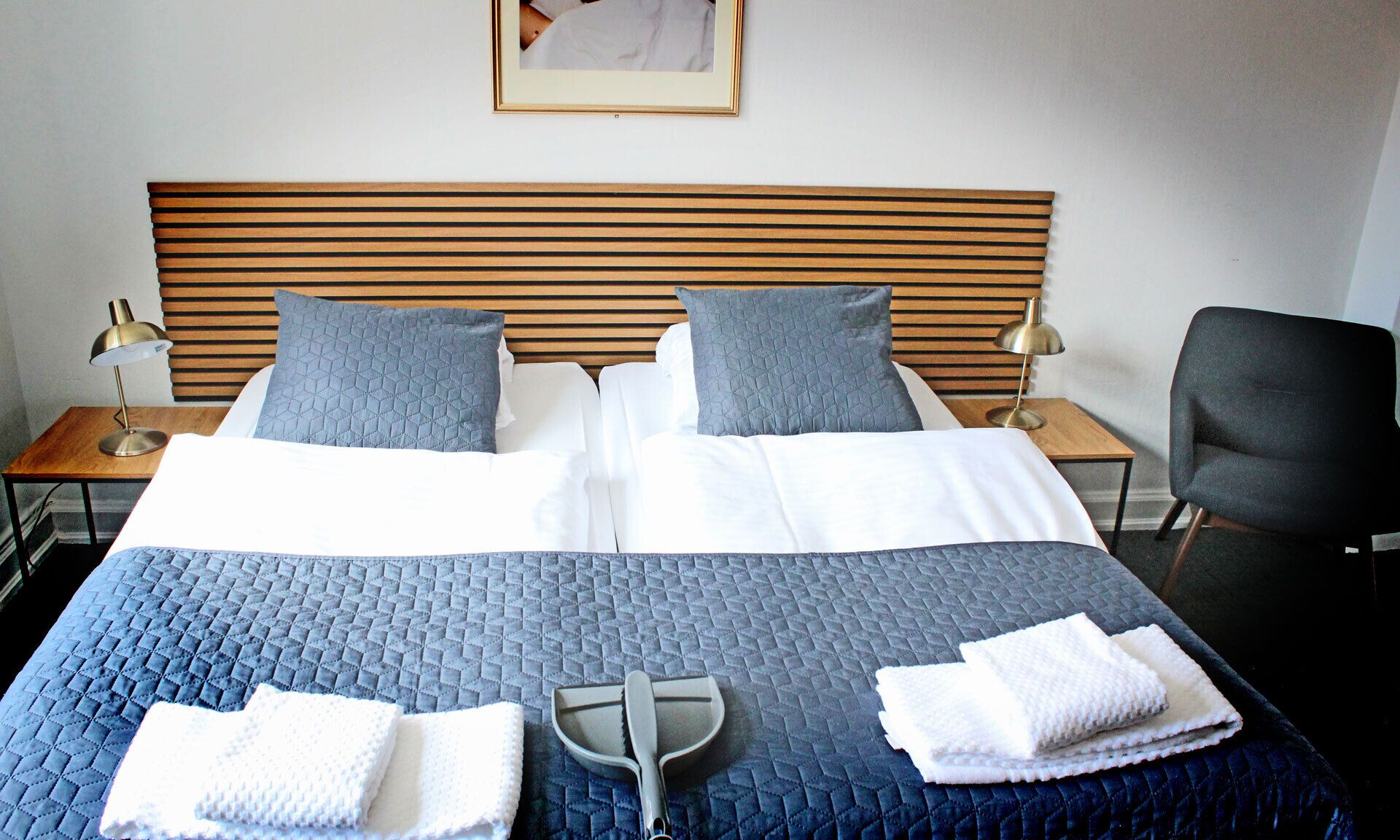 Un lit double avec une couette bleue, deux oreillers, une tête de lit en bois, une table de chevet avec une lampe, une chaise et un tableau au mur.