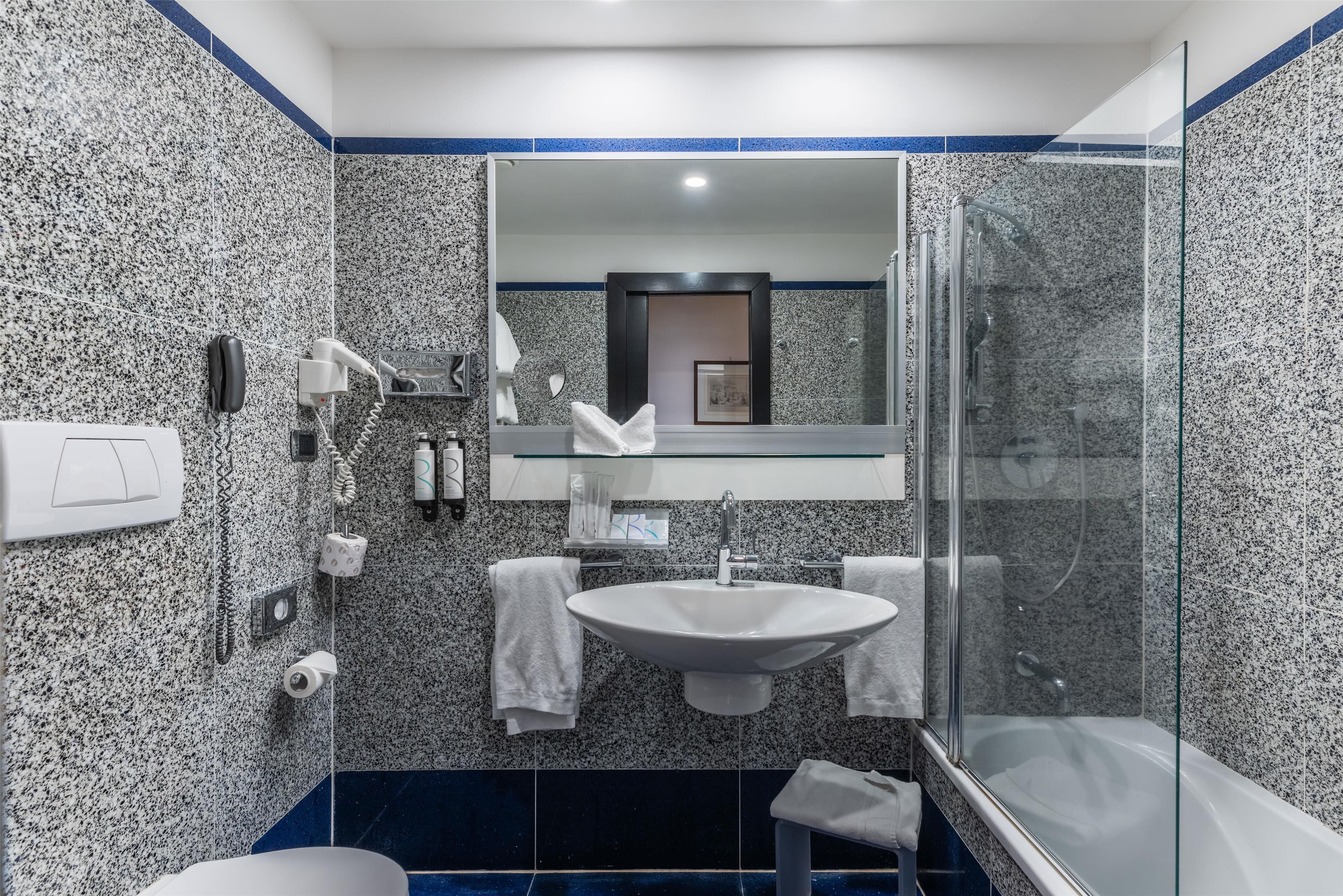 A modern bathroom with a granite backsplash, a large mirror, a sink, a glass-enclosed shower, and a toilet.