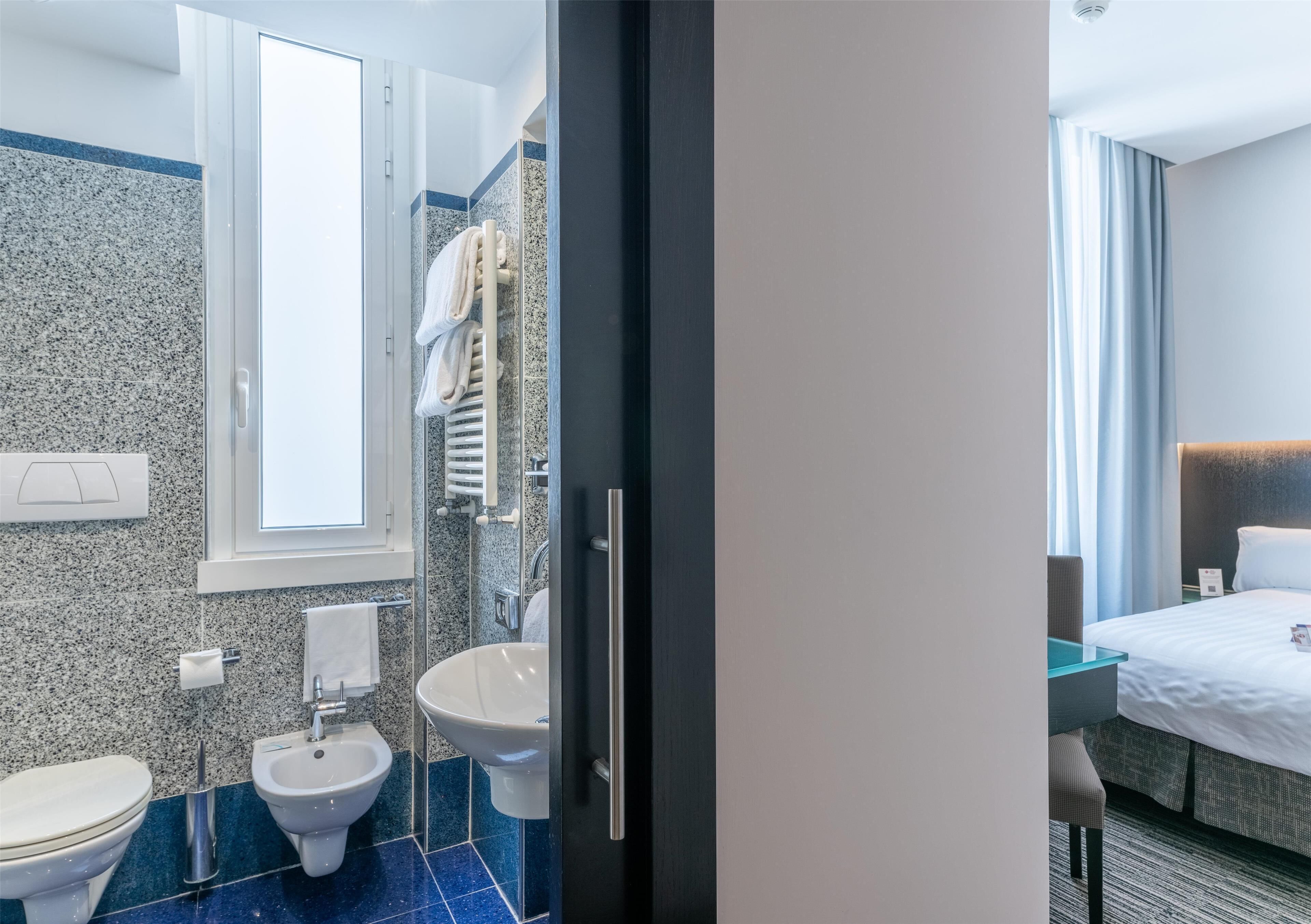 Un bagno moderno in hotel con bidet, WC e lavandino. Si vede una finestra che lascia entrare luce naturale e offre vista sulla stanza.