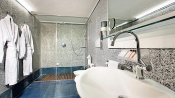 Un baño moderno con un lavamanos grande, cabina de ducha con mampara de vidrio y un perchero con batas blancas.