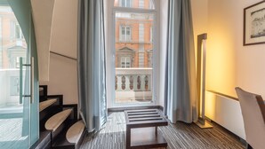 Une chambre d’hôtel avec vue sur un immeuble par la fenêtre, un banc en bois et une lampe sur pied.
