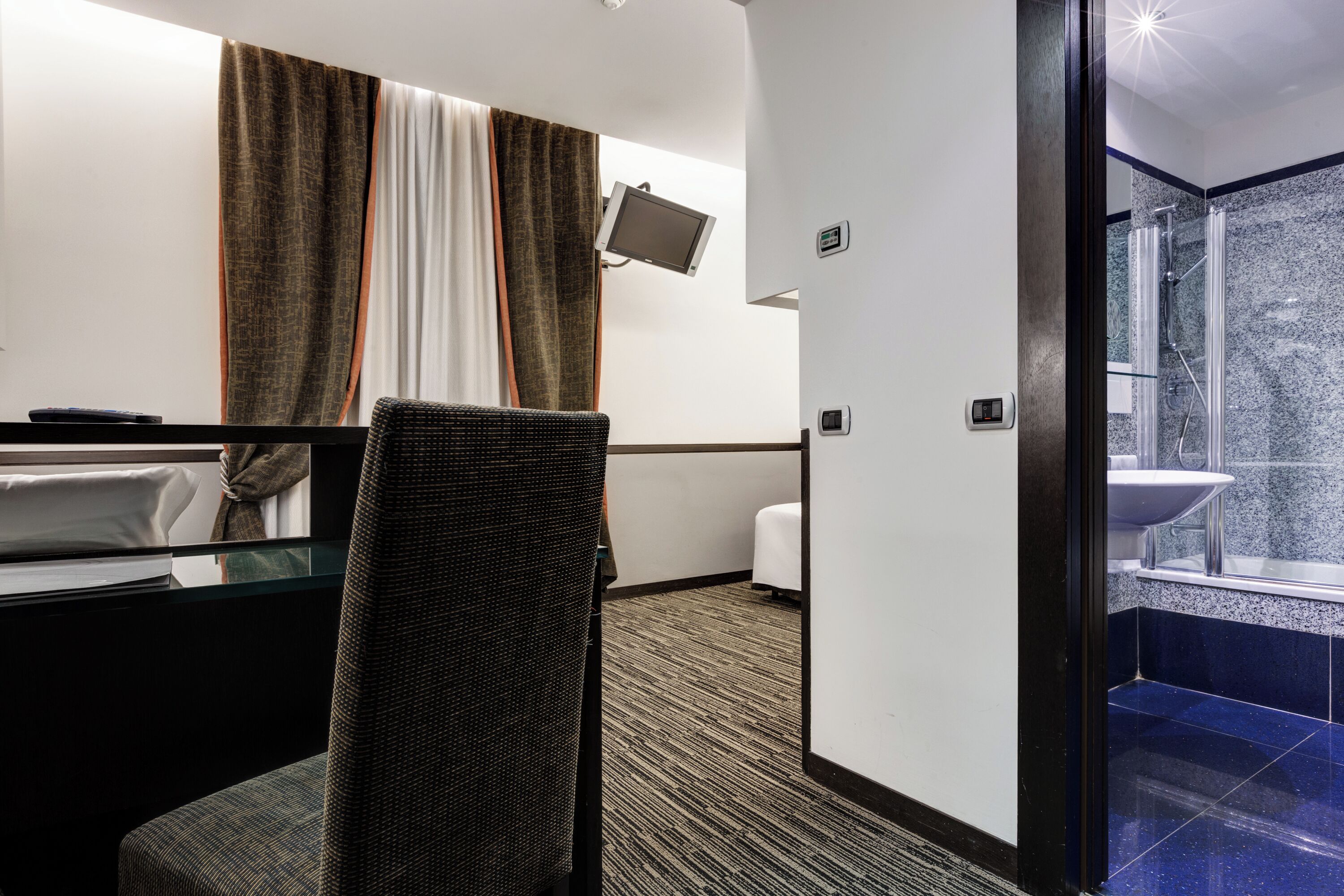 A hotel room with a bed, a desk, a chair, a TV, and a bathroom with a shower and sink.