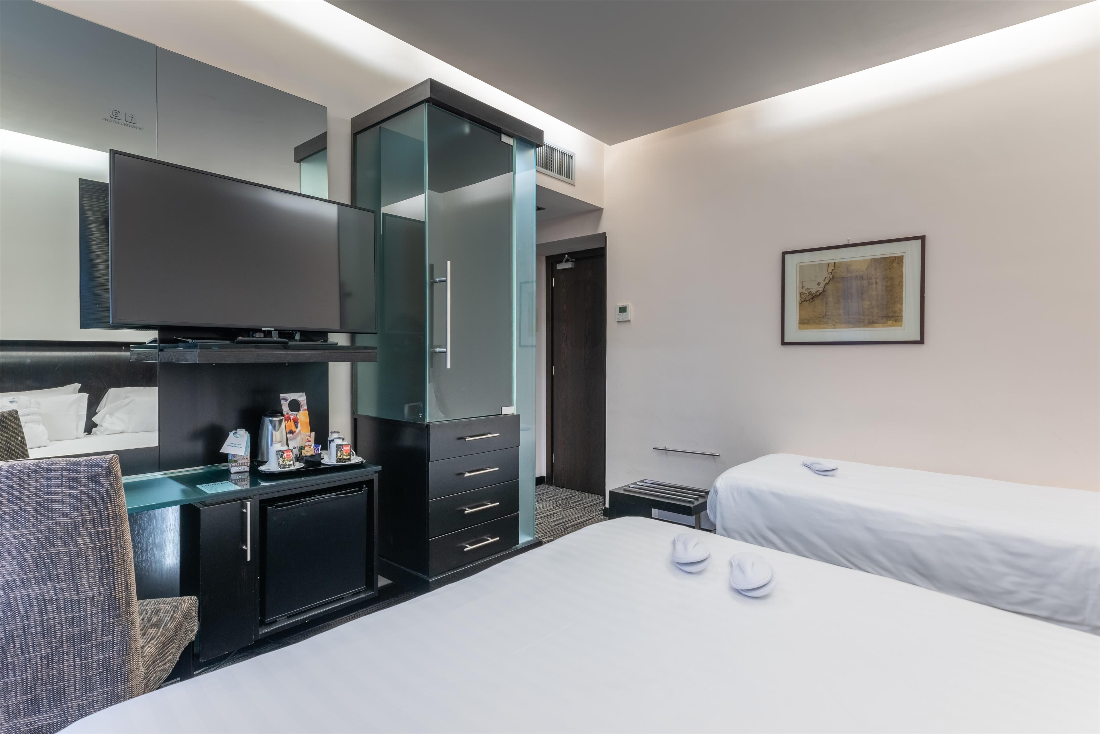 A hotel room with a bed, a desk with a flat-screen TV, a minibar, a chair, and a door leading to another room.