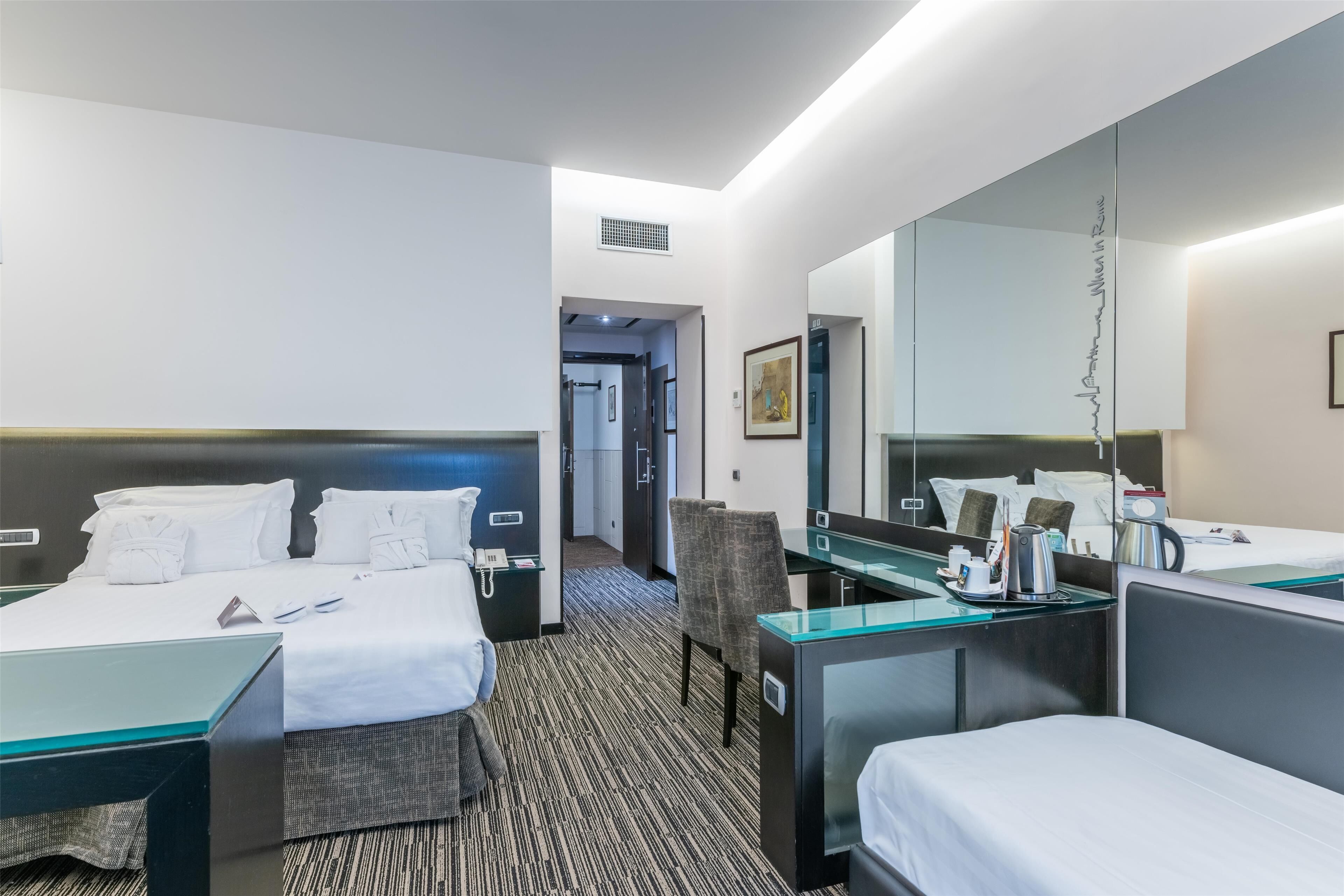 A hotel room with two beds, a desk, a chair, a large mirror, and a coffee maker.