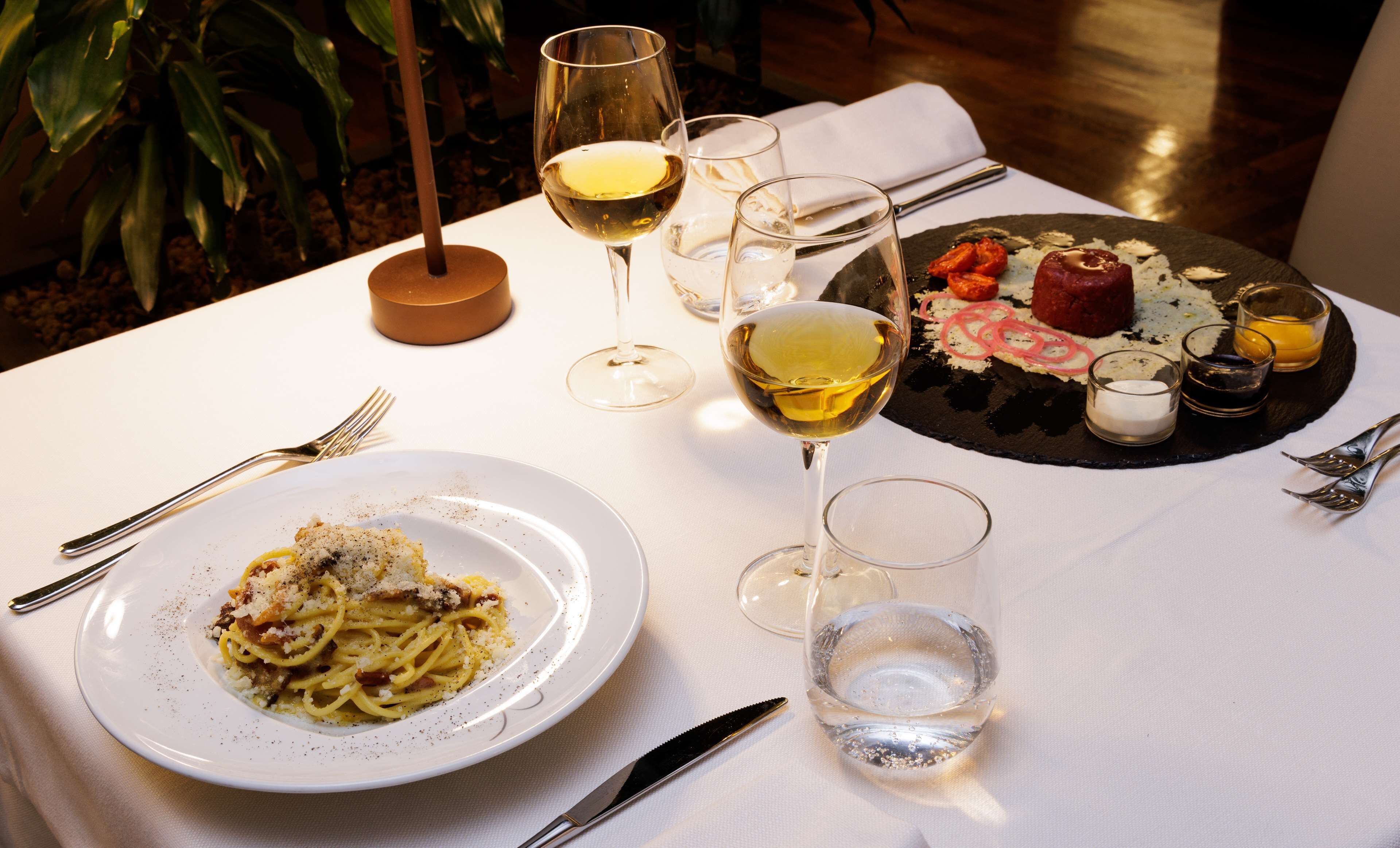 Een eettafel met pasta, een vleeswarenbord, glazen witte wijn en bestek.