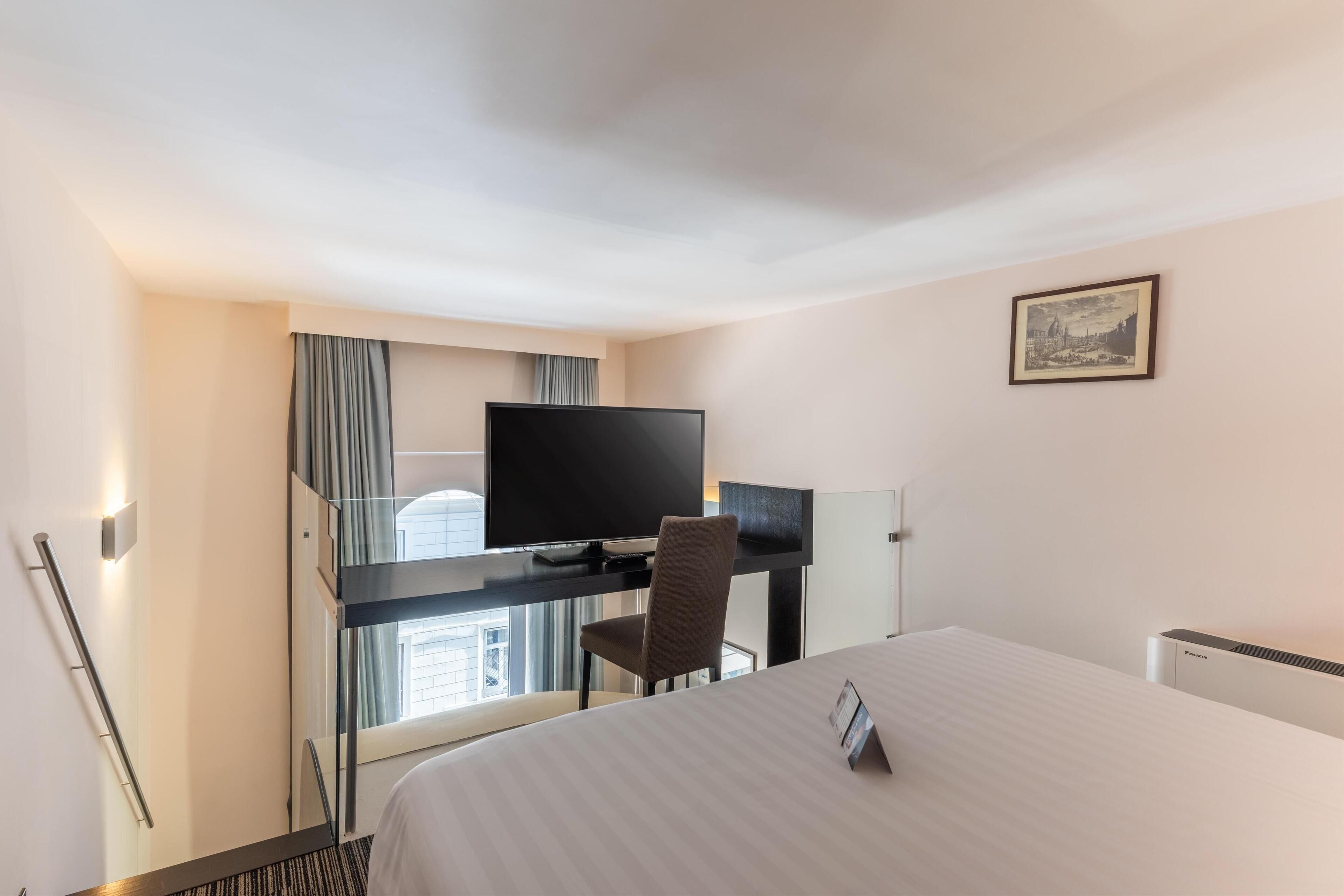 A hotel room with a bed, a desk with a TV, a chair, and a window with curtains.