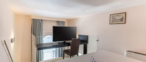 A hotel room with a bed, a desk with a TV, a chair, and a window with curtains.
