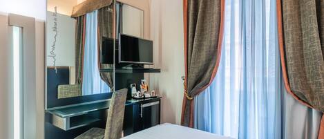 A hotel room with a bed, a desk with a flat-screen TV, a chair, and a minibar.