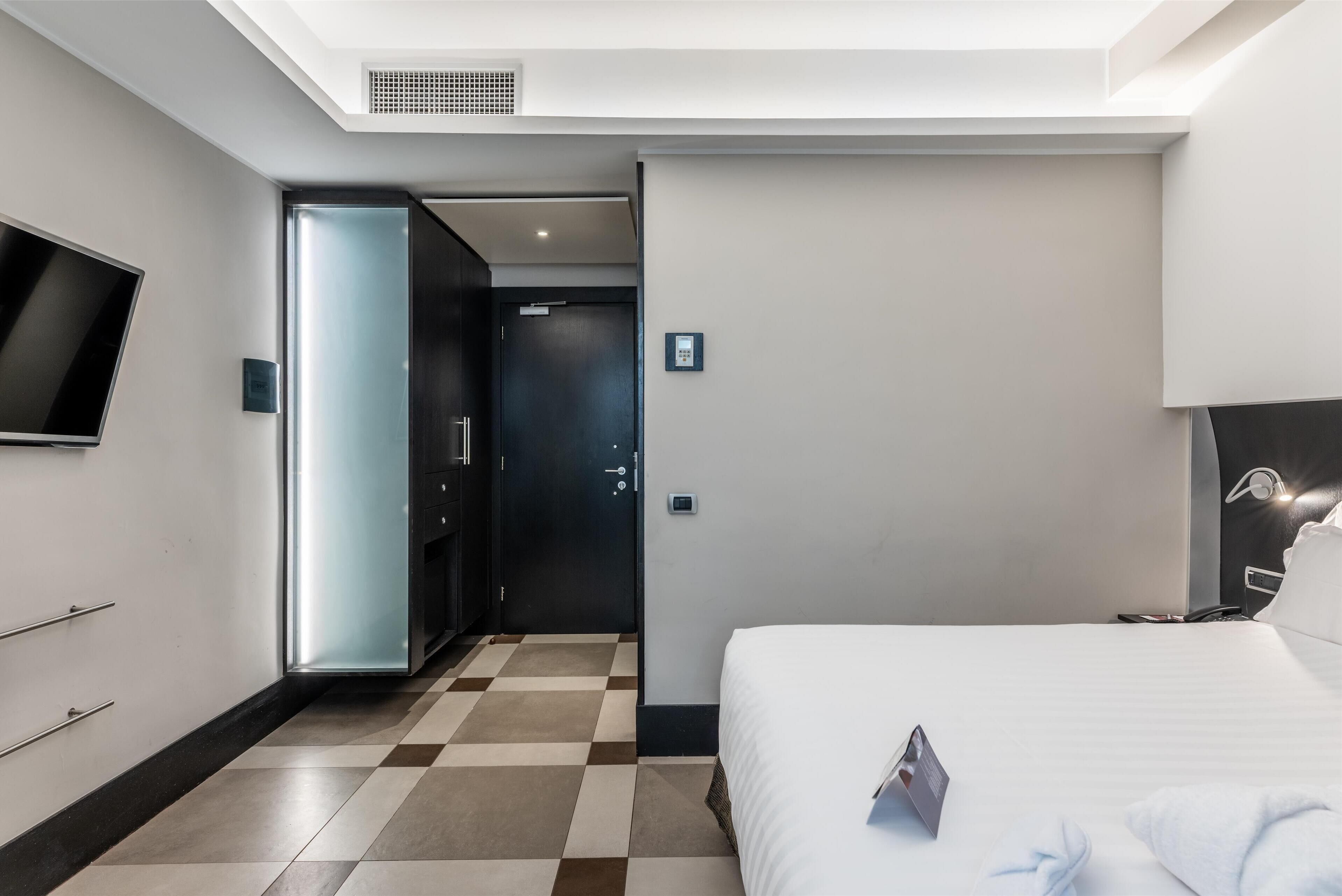 A modern hotel room with a large bed, a flat-screen TV mounted on the wall, a door leading to a bathroom, and a check-in kiosk.