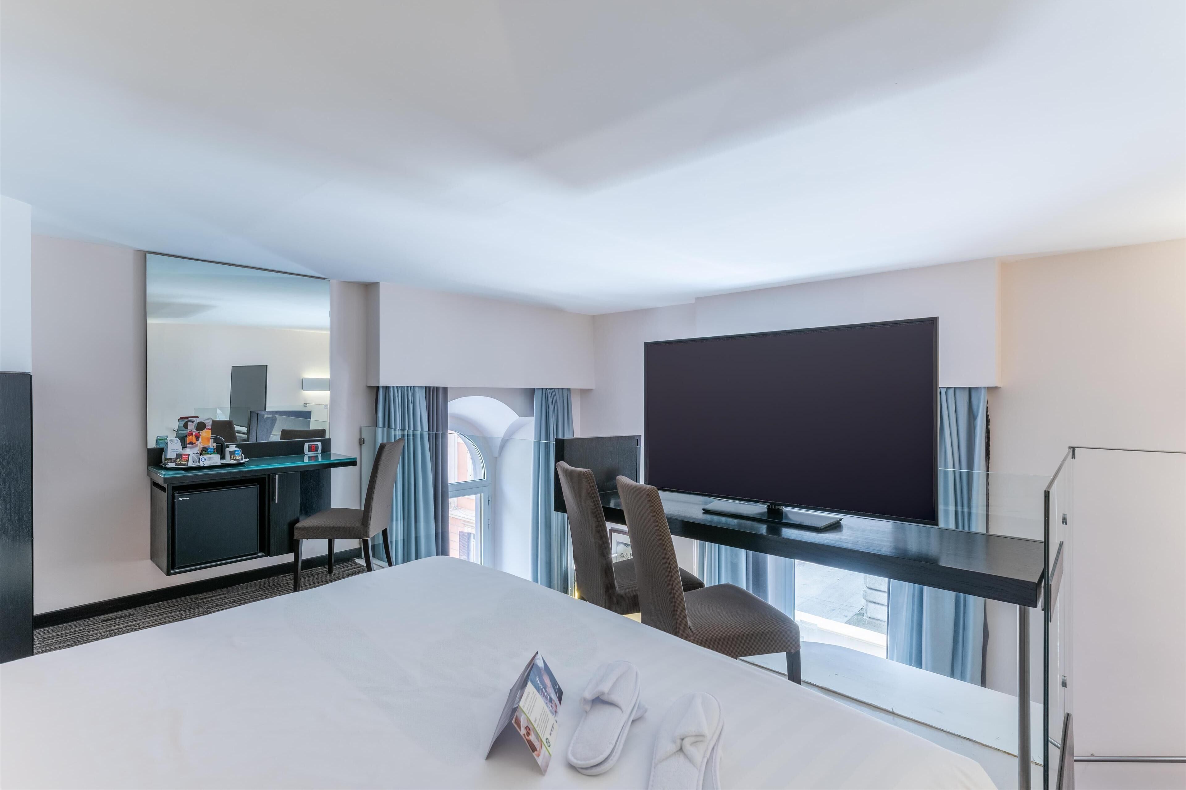 Habitación de hotel con una cama, un escritorio con computadora, televisor y ventana con cortinas.