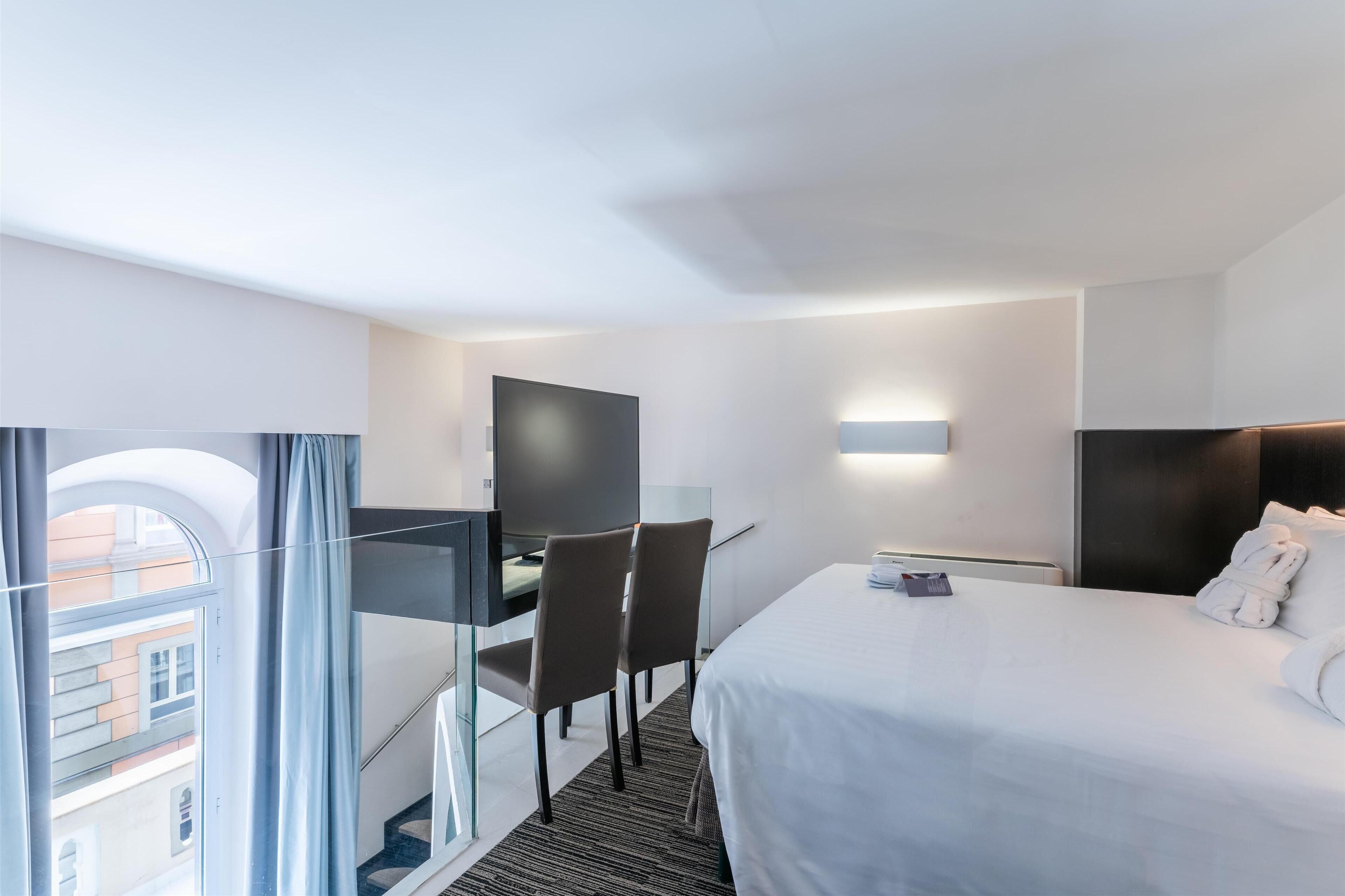 A hotel room with a bed, desk, chairs, a TV, and a window with curtains.