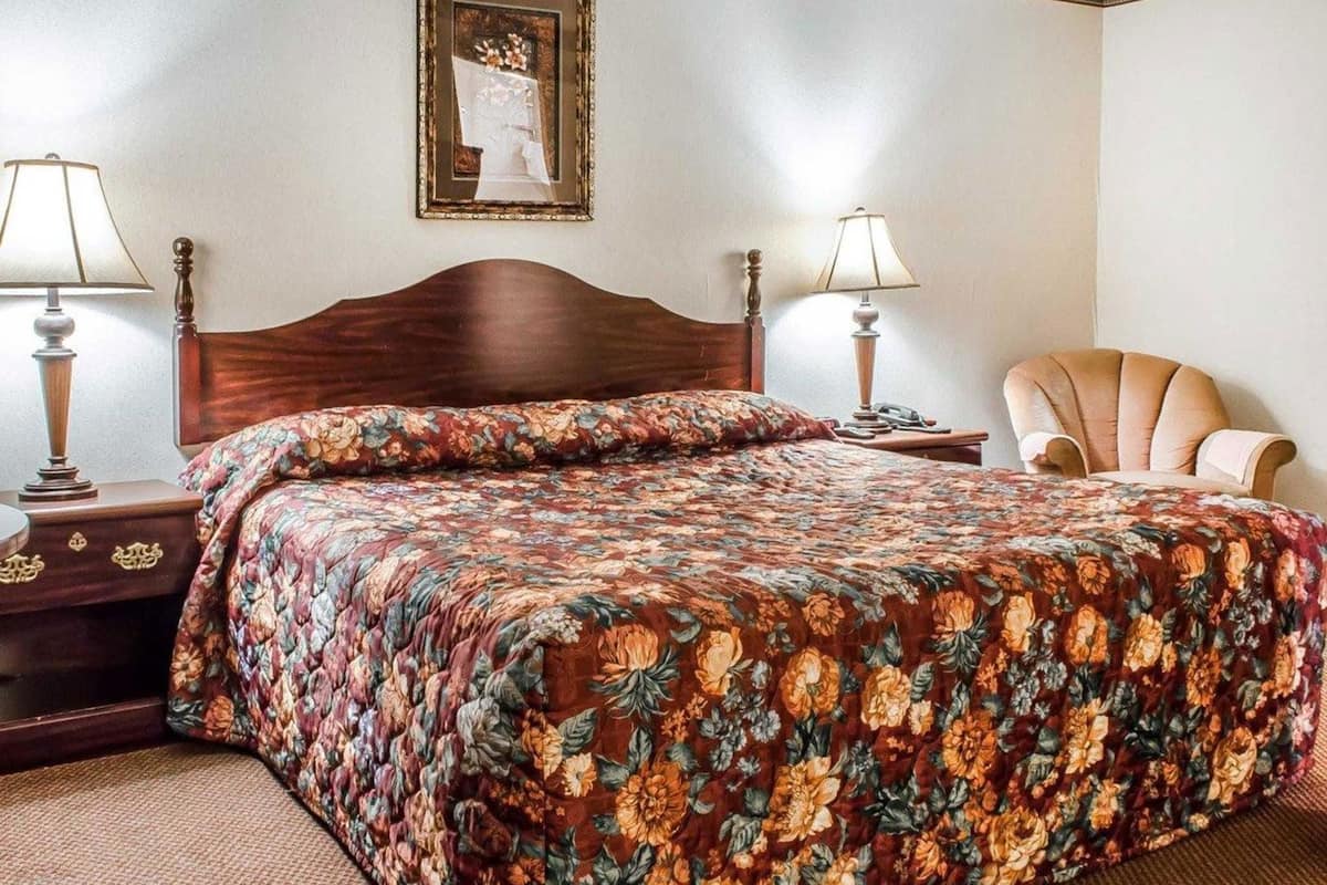 Een hotelkamer met een houten bed, een beddengoed met bloemenprint, twee bedlampen, een stoel en een lijst aan de muur.