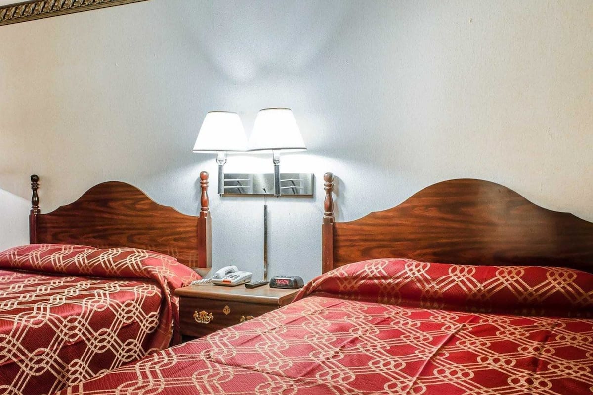 Hotelkamer met een houten hoofdeinde, twee bedden met rode, gemêleerde beddengoed, een nachtkastje met telefoon en klok, en twee wandlampen.