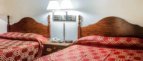 A hotel room with a wooden headboard, two beds with red patterned bedding, a nightstand with a phone and a clock, and two wall-mounted lamps.