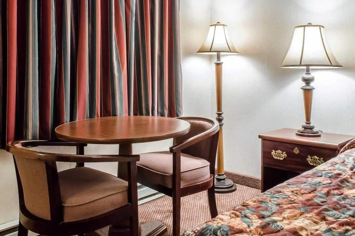 Een hotelkamer met een bed, een ronde tafel met twee stoelen, een nachtkastje met een lamp en een raam met gordijnen.