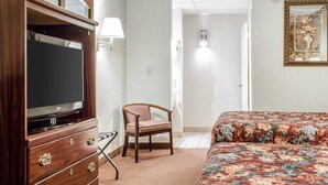 Quarto de hotel com cômoda de madeira, televisão, colcha estampada com flores, uma cadeira e abajur.