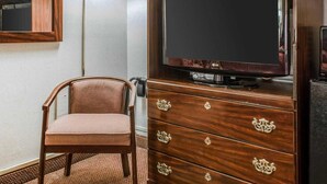 Quarto de hotel com cómoda de madeira, televisão, cadeira e candeeiro fixo na parede.