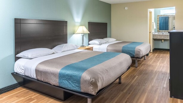 A hotel room with two beds, a nightstand with a lamp, a bathroom visible through an open door, and a wooden floor. - Motel 6 Kenly, NC (Kenly)