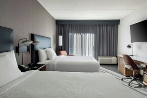 A hotel room with two beds, a desk, a chair, a TV, and a large window with curtains.