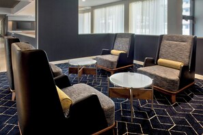 A modern hotel lobby with a geometric patterned carpet, comfortable seating with cushions, and a large window offering a view of the city.