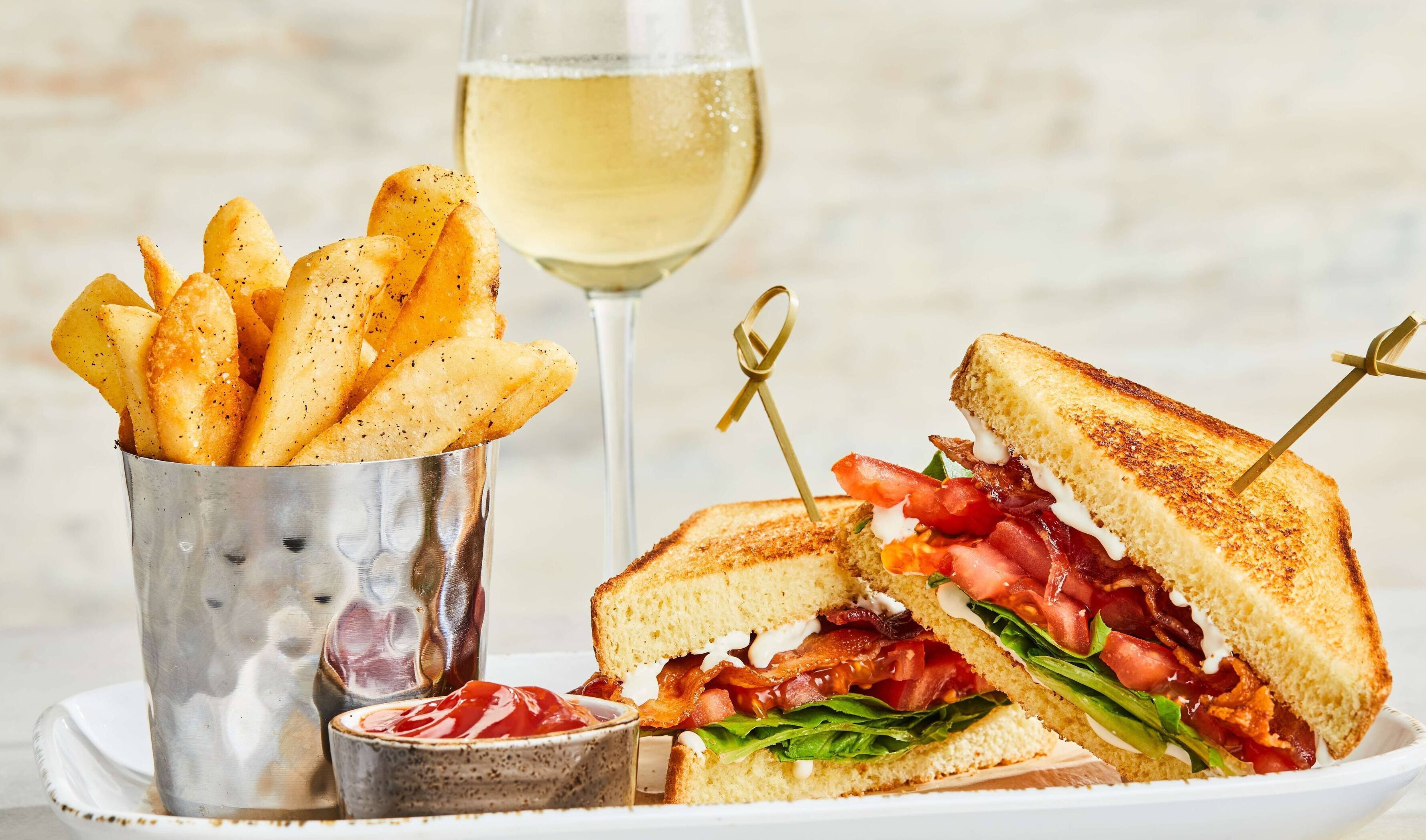 Um sanduíche com bacon, tomate e alface, um copo de vinho branco e um prato de batatas fritas.
