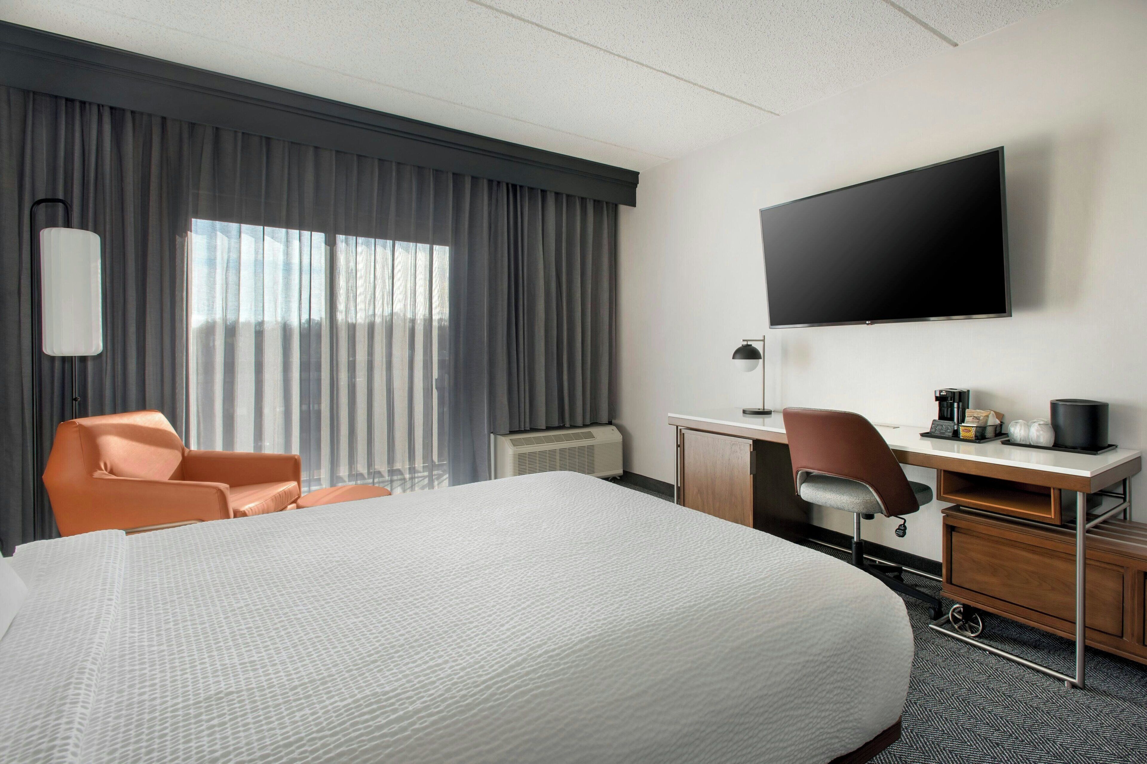A hotel room with a large bed, a desk with a chair, a flat-screen TV, and a window with curtains.