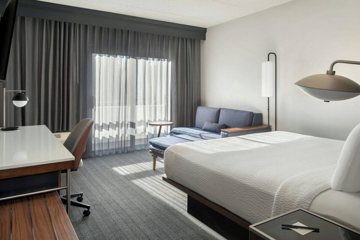A hotel room with a bed, a desk, a chair, a lamp, and a window with curtains.
