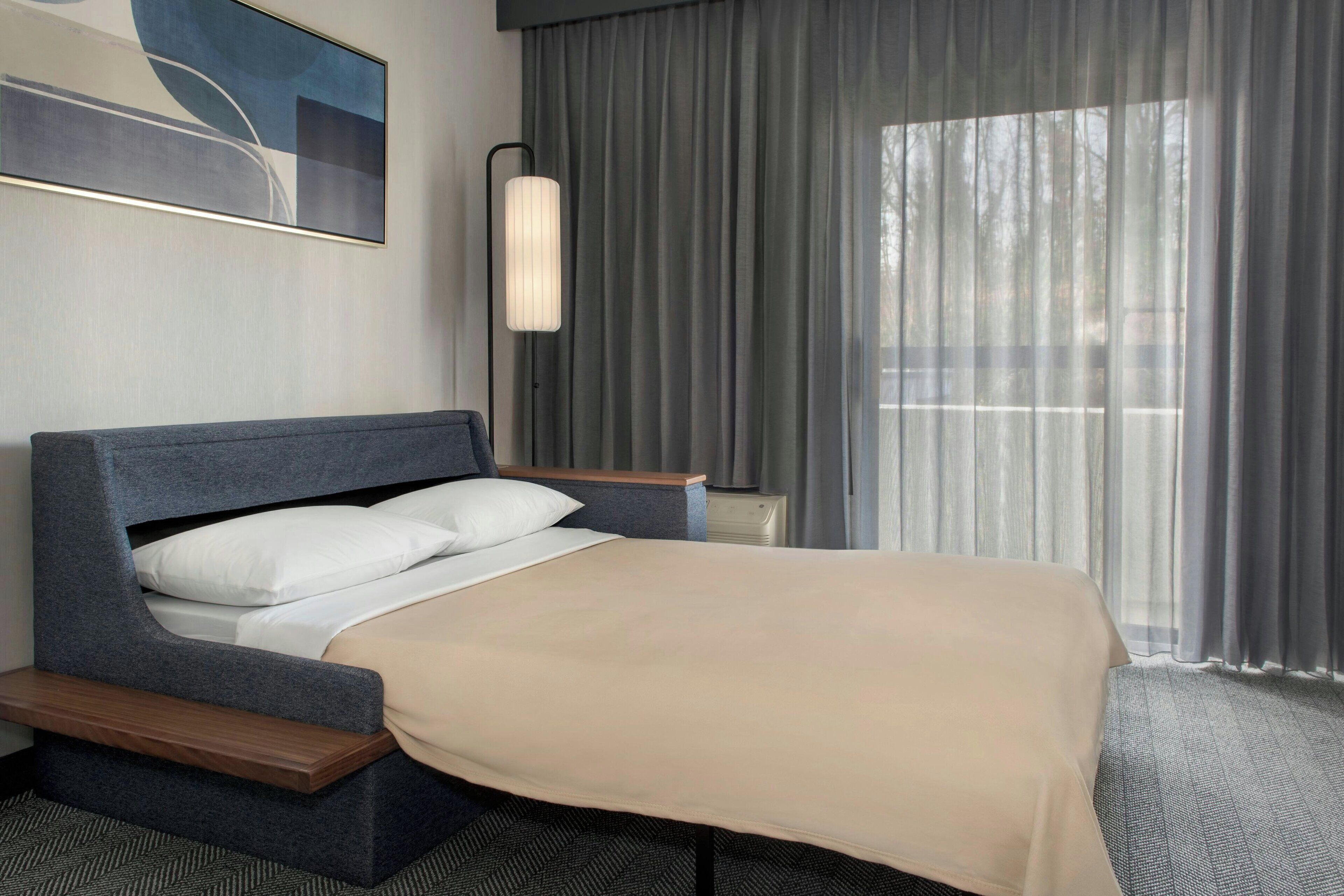 A modern hotel room with a bed, a bedside table, a lamp, and a large window with curtains.