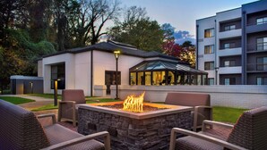 A patio area with a stone fire pit and wicker chairs, surrounded by a building with a glass enclosure and a white exterior wall. - Courtyard by Marriott Rye (Rye Brook)