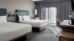 A hotel room with two beds, a desk, a chair, a TV, and a large window with curtains. - Courtyard by Marriott Rye (Rye Brook)