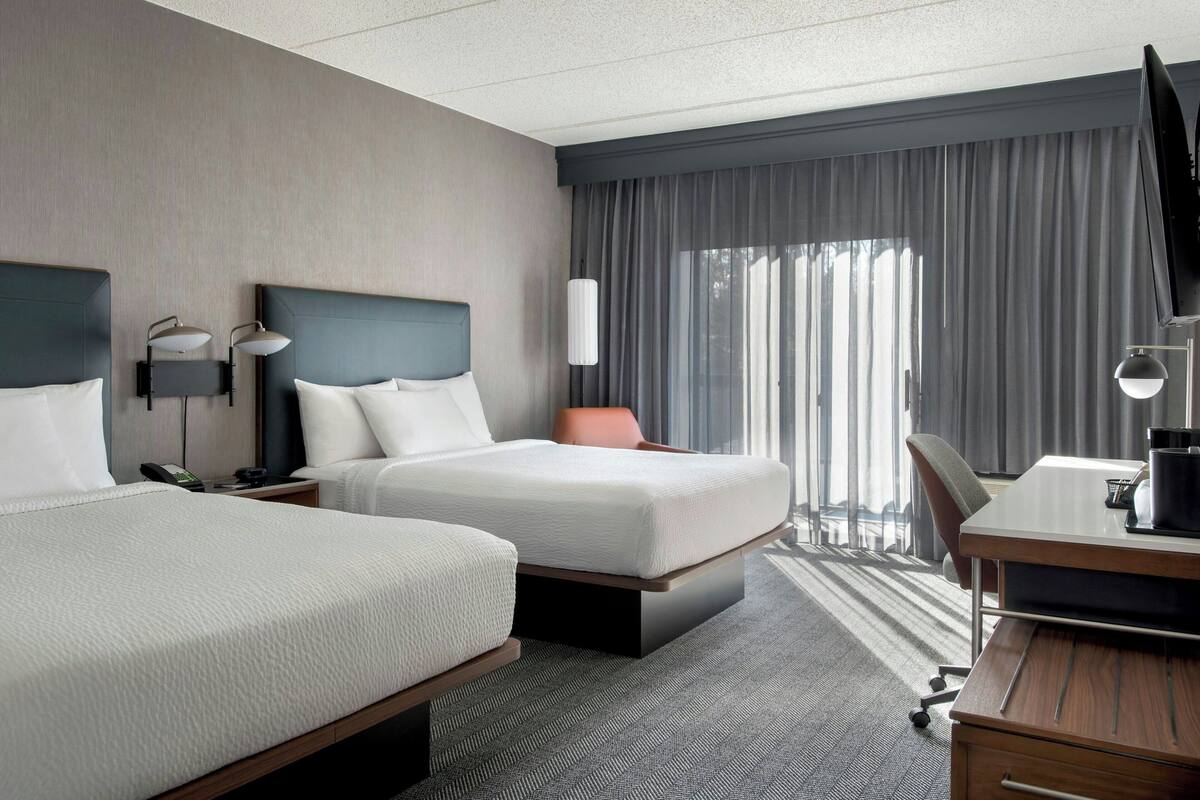 A hotel room with two beds, a desk, a chair, a TV, and a large window with curtains.