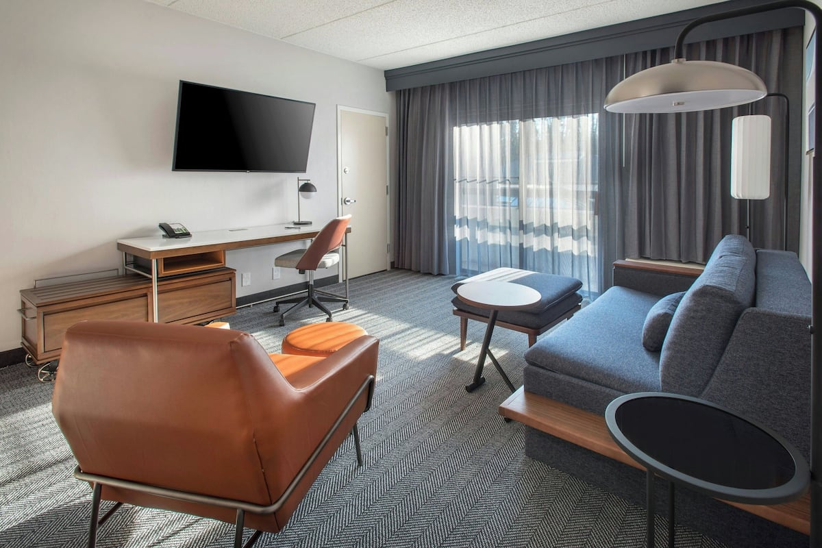A hotel room with a flat-screen TV, a desk, a brown leather chair, a grey sofa, a small round table, and a large window with curtains.