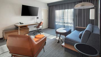 Habitación de hotel con televisor de pantalla plana, escritorio, sillón de cuero marrón, sofá gris, mesita redonda y ventana grande con cortinas.