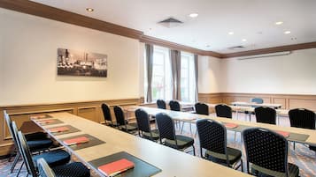 Et mødelokale med et langt bord, flere stole, et projektorskærm og et stort vindue med gardiner.