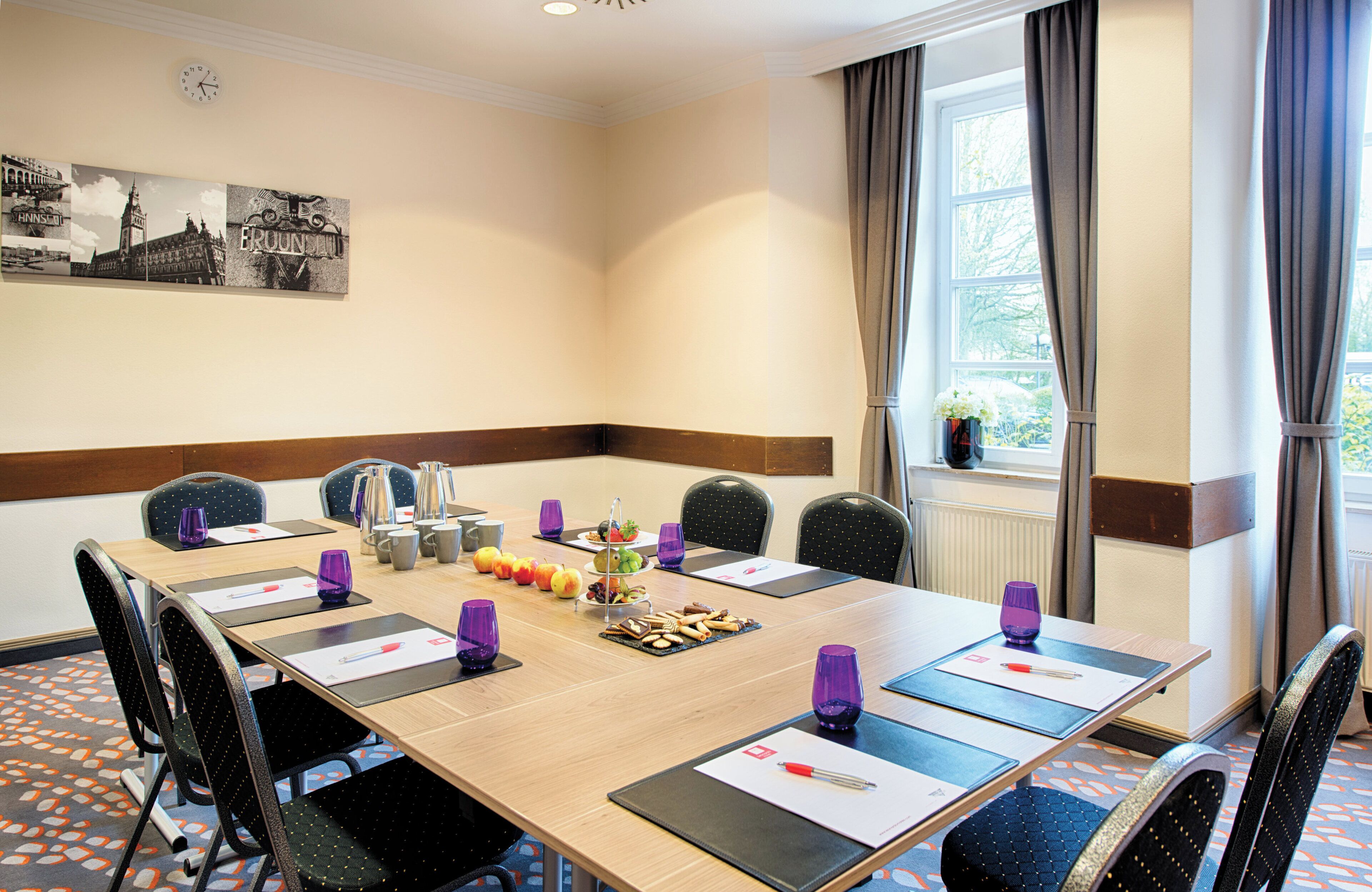 Une salle de conférence avec une longue table, des chaises, une horloge et une fenêtre avec des rideaux.