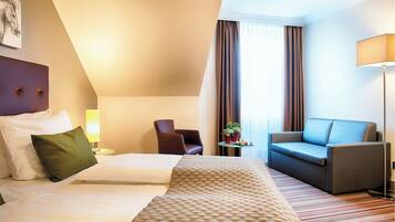 A hotel room with a bed, a sofa, a chair, a lamp, and a window with curtains.