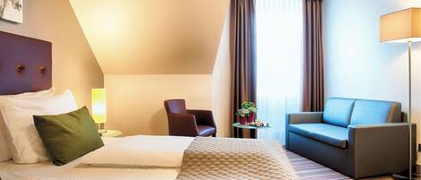 Una habitación de hotel con cama, sofá, silla, lámpara y ventana con cortinas.