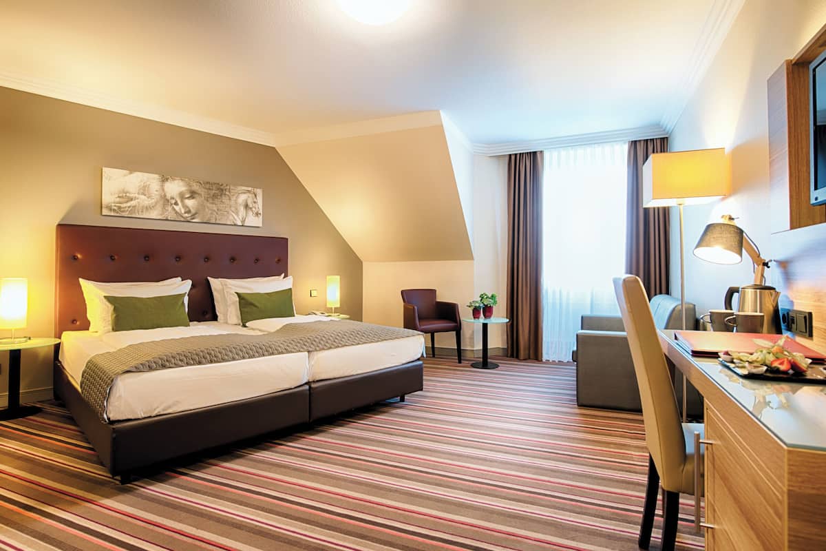 A hotel room with a large bed, a desk with a lamp, a chair, and a small table with plants.