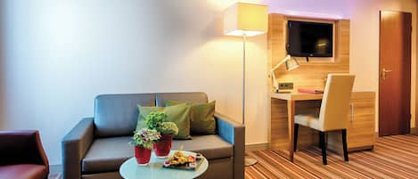 A hotel room with a sofa, a small table with a plant and snacks, a desk with a chair, a TV mounted on the wall, and a door.