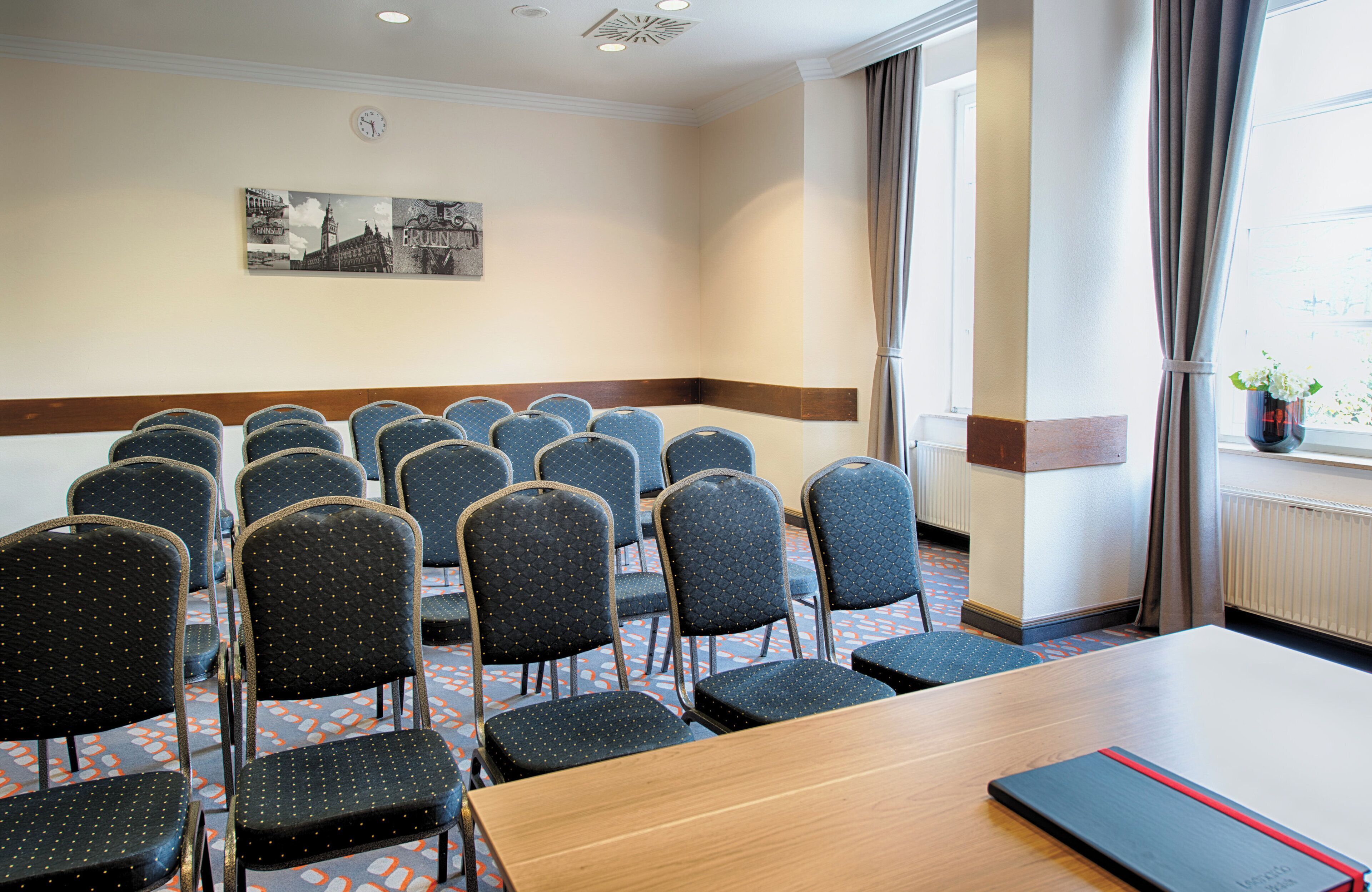 Una sala conferenze con file di sedie, un lungo tavolo, un orologio e una finestra con tende.