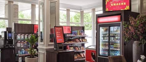 Un bar de aperitivos en hotel con un quiosco de autoservicio, mostrador refrigerado para bebidas y una zona de estar con una planta en maceta.