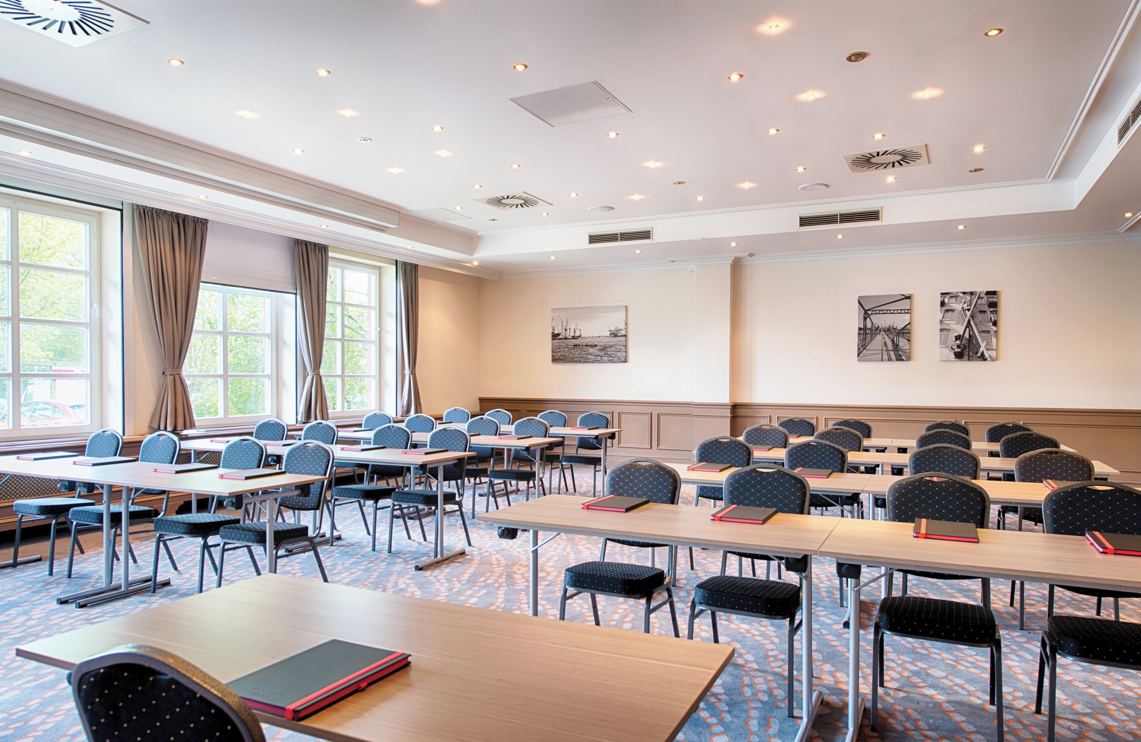 A conference room with multiple tables and chairs, large windows with curtains, and ceiling lights.