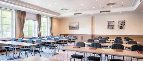 A conference room with multiple tables and chairs, large windows with curtains, and ceiling lights.
