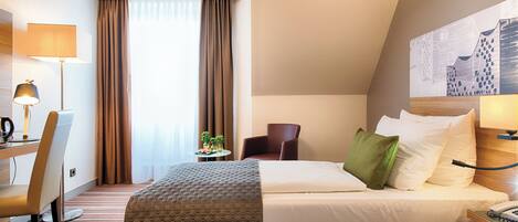 A hotel room with a bed, a desk with a lamp, a chair, and a small table with a plant.
