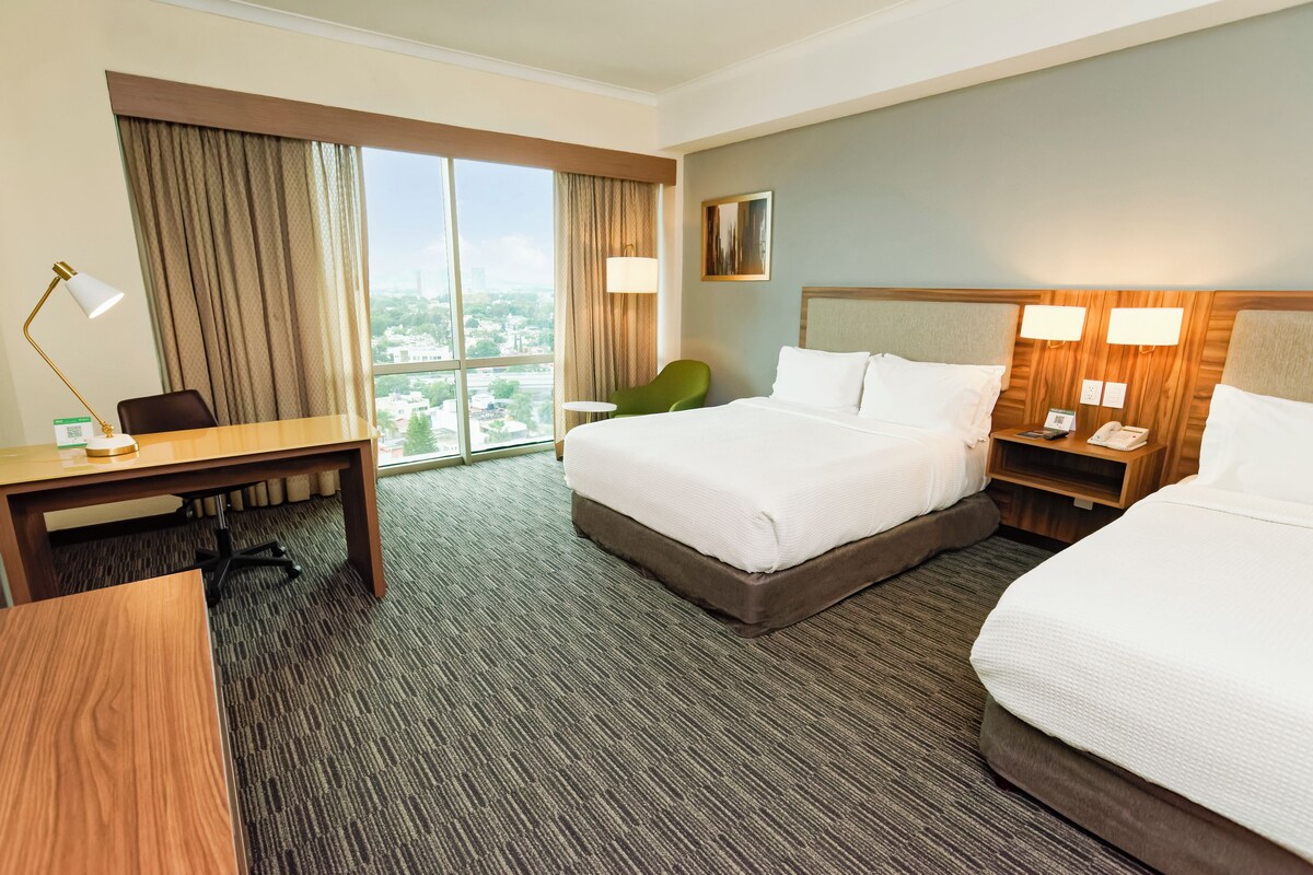 A hotel room with a large bed, a desk, a chair, a nightstand, and a view of the city.