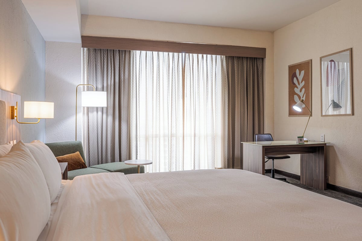 A hotel room with a large bed, a desk, a chair, a small table, and a window with curtains.