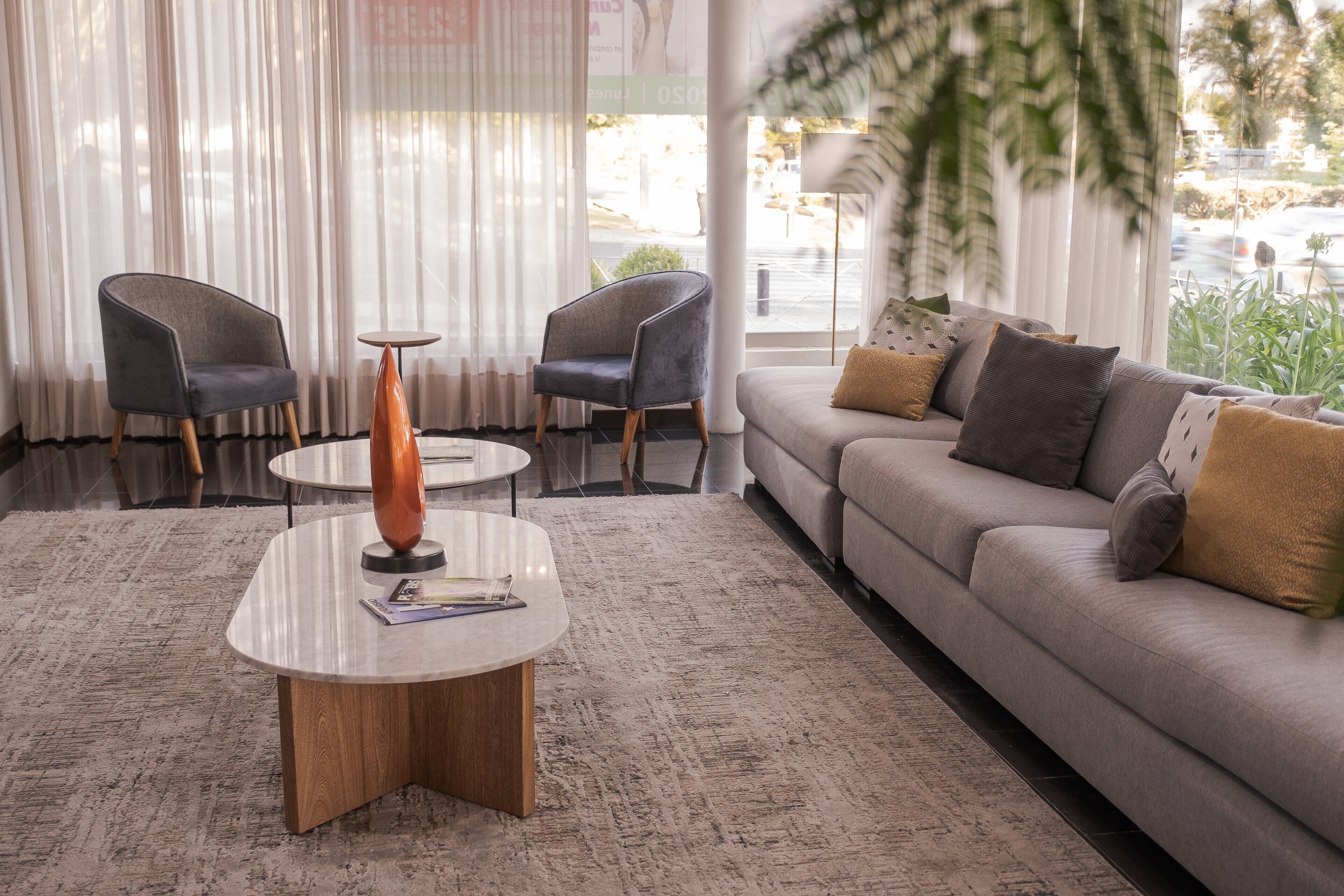 A modern living room with a sofa, two armchairs, a coffee table, and a view of the street through large windows.